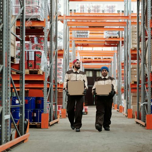 Two workers carrying boxes walk between metal shelves in a spacious warehouse.